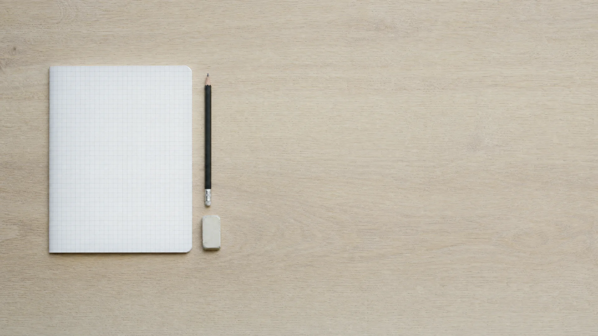 Overhead view of a pale oak table with a closed squared-grid exercise book, a sharpened pencil and a small eraser, with wide empty space beside them.